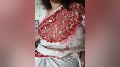 Video of a woman in a red and white saree with intricate embroidery, seated against a blurred background, showcasing her midriff and upper body.