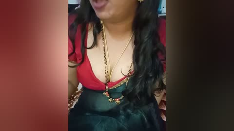 A video of a South Asian woman with long, wavy black hair, wearing a deep red blouse, black sari, gold necklace, and earring, sitting on a couch.