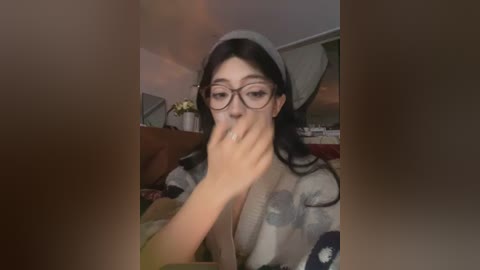 Video of a young woman with long black hair, wearing glasses and a grey hat, holding a phone to her ear, seated in a dimly lit room with a green plant and a wooden table in the background.