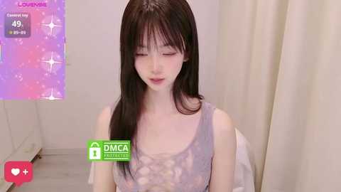 A video of a fair-skinned Asian woman with long, straight black hair and bangs, wearing a light gray, floral-patterned tank top, seated indoors. The background features a soft-focus, cream-colored curtain.