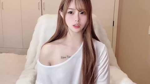 Media: Video of a young Asian woman with long, straight brown hair, fair skin, and large breasts, wearing a white off-shoulder top, sitting on a white bed, with white closet doors in the background.