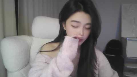 A video of an East Asian woman with long black hair, wearing a light pink fuzzy robe, sitting on a white chair, looking down with a fur-covered object in her mouth.
