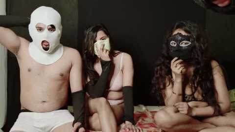Video of three individuals in a dimly lit room: a shirtless man in a white mask, a woman with long hair in a green mask, and a woman with long hair in a black mask.