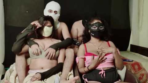 Video of four women in masks, lingerie, and gloves, seated on a couch; one woman is blindfolded. Background shows a dark room with a white curtain.