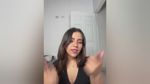 Video of a young woman with medium skin tone and long brown hair, wearing a black V-neck dress, standing in a white-walled room with a white door. She has her hands up, appearing to be dancing.