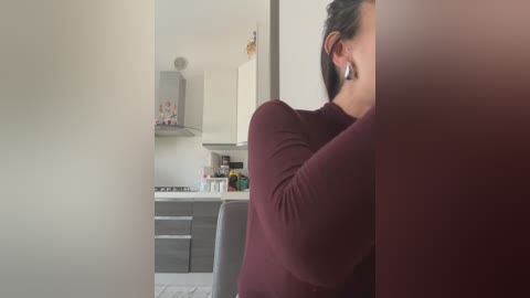 A video of a woman with medium skin tone and dark hair, wearing a burgundy sweater, hiding behind a doorway, in a modern kitchen with white cabinets, gray countertops, and a stove.