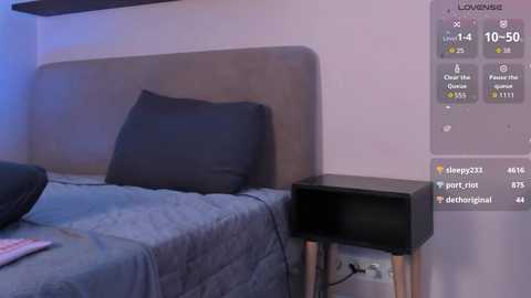 Video of a minimalist bedroom with a gray bed, black pillow, and a wooden bedside table. The room has a white wall with a virtual assistant screen displaying controls and settings.