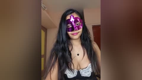 A video of a young woman with long, dark hair and medium skin tone, wearing a purple Venetian mask and a black lace-trimmed bra, standing in a dimly lit room with beige walls.