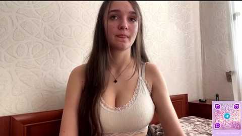 Media: Video of a young, fair-skinned woman with long, straight brown hair, wearing a white lace-trimmed camisole, sitting on a bed with patterned bedding, in a room with textured wallpaper.