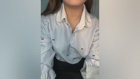 Video of a woman wearing a blue and white striped blouse with a white collar, black skirt, and visible chest area. Background is plain light gray.