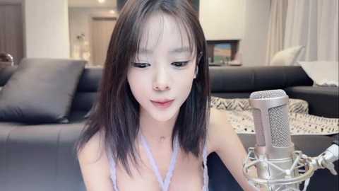 Video of an Asian woman with straight, shoulder-length black hair, wearing a light purple spaghetti strap top, singing into a silver microphone. She's in a modern living room with black leather sofas and white curtains.
