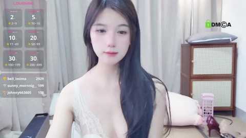A video of an East Asian woman with long, straight black hair, wearing a lacy white bra, sitting at a desk with a digital clock, a remote, and a phone. The room is lightly lit with beige curtains.