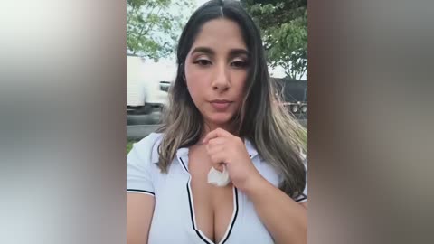 A video of a Latina woman with medium skin tone and long, dark hair, wearing a low-cut white polo shirt, holding a white tissue near her cleavage, outdoors with blurred trees and a white truck in the background.
