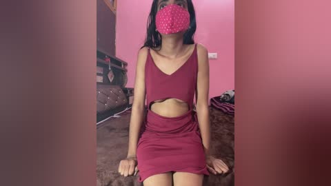 Media: Video of a slender, dark-skinned woman with long black hair, wearing a red crop top and skirt, sitting on a bed with a pink mask covering her face, in a pink room.