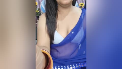 A video of a woman with medium skin tone, wearing a blue saree with a white bra underneath, holding a glass of water. Sunflowers and a vase are in the background.