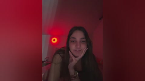 A dimly lit video of a young woman with long dark hair, smiling, with one hand on her face, in a red-lit room.
