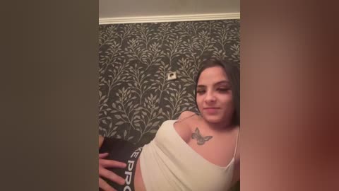A video of a smiling, fair-skinned woman with brown hair, wearing a white tank top and a black sports bra, holding a phone, in a room with dark floral wallpaper.