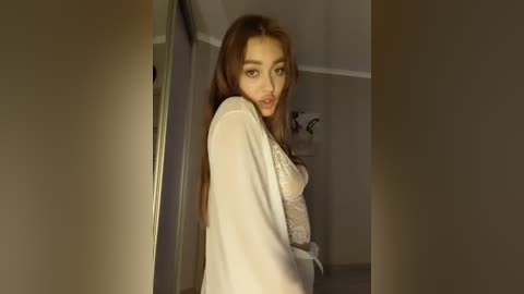 Video of a young woman with long, straight brown hair, wearing a sheer white lace robe, standing in a dimly lit room, with beige walls and a framed picture in the background.