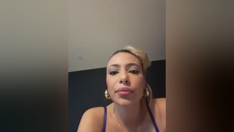 Media: A video of a woman with fair skin, blonde hair in a bun, and large gold hoop earrings, wearing a dark blue top. She has a neutral expression and is looking directly at the camera against a plain, dark background.