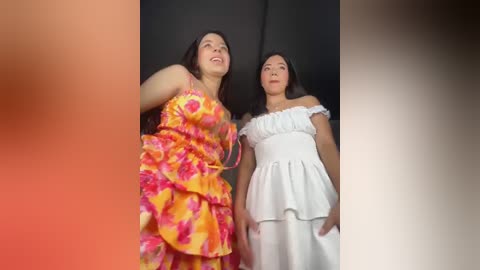 Video of two women, one in a vibrant yellow floral dress, the other in a white off-shoulder dress, standing close together against a black backdrop.