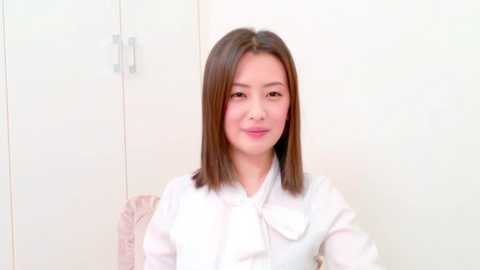 Video of a young East Asian woman with straight, shoulder-length brown hair, wearing a white button-up shirt, sitting in front of a plain white wall with a beige cushion.