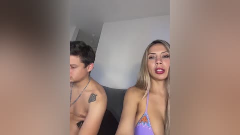 A video of a topless shirtless man with a tattoo and a blonde woman in a purple bikini, both facing away from the camera in a dimly-lit room.