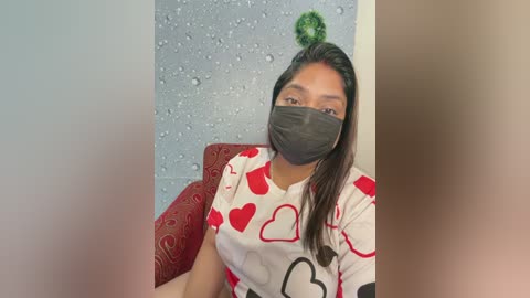 Video of a young woman with medium brown skin and long black hair, wearing a gray face mask, white t-shirt with red heart designs, and red-patterned cushion, against a speckled blue wall.