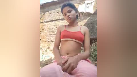 Video of a young woman with dark hair and light skin, wearing a red bra and pink pants, sitting outdoors against a rustic stone wall.