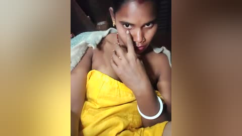 A video of a young woman with dark skin, wearing a yellow saree, sitting on a bed, making a peace sign with her right hand. She has a nose ring and a bracelet on her wrist.
