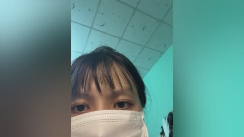 Video of a young Asian woman with short dark hair, wearing a white face mask, looking up. The background features a turquoise wall and a grid-patterned ceiling.