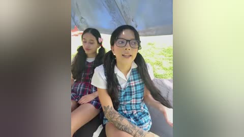 Media: Video of two young Asian girls in school uniforms, one with glasses, sitting side by side in a car, sunlight filtering in.