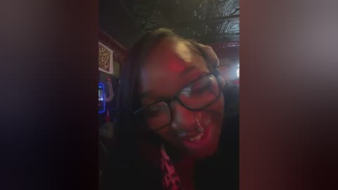 Video of a smiling Black woman with braided hair, wearing glasses and a pink shirt, in a dimly lit room with red lighting.