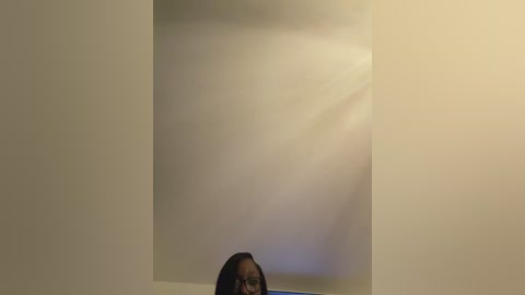 Video of a blurred person with dark hair and glasses, standing in a dimly lit room with beige walls and a white ceiling. The image has a soft, muted color palette.