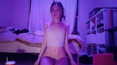 Video of a slender, petite Asian woman with dark hair in a messy bun, wearing a white ribbed tank top and matching panties, sitting on a carpeted floor in a dimly lit bedroom with purple lighting.