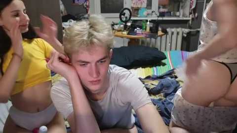 Video of a young, pale-skinned, platinum-haired boy in a white T-shirt, lying on a bed with a messy room in the background. Two girls in bikinis and lingerie are nearby.