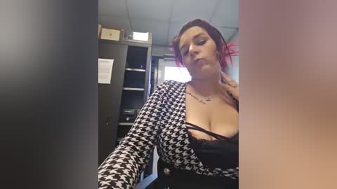 A video of a woman with red hair, wearing a black and white houndstooth jacket, revealing a black lace bra, standing in a cluttered office with shelves and a window.