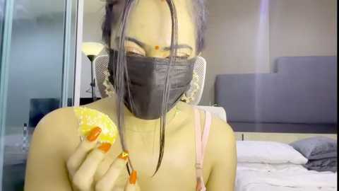 Video of a woman with light skin and dark hair in braids, wearing a black face mask and a pink top, holding a yellow slice of pineapple, in a modern bedroom with a bed and lamp.