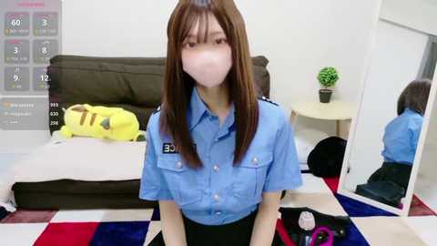 Video of a young Asian woman with straight brown hair, wearing a blue police uniform and face mask, sitting on a multi-colored rug in a sparsely furnished room with a bed, potted plant, and mirror.