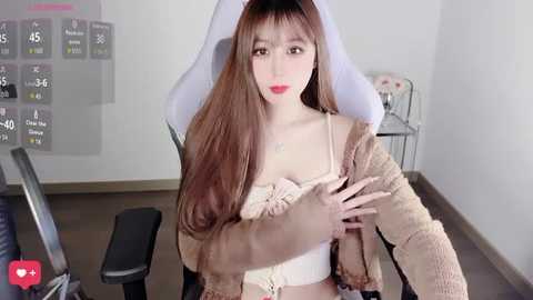 A video of a fair-skinned woman with long brown hair, wearing a beige jacket, sitting in a white gaming chair. The background shows a simple room with a desk and a grayboard.