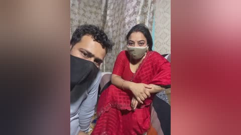 Video of a man with short black hair in a blue shirt and a woman in a red checkered saree with a mask, seated indoors with patterned wallpaper.