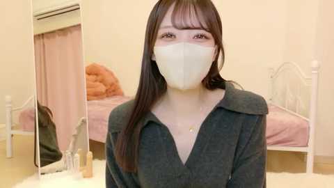 Video of a young Asian woman with long, straight black hair, wearing a gray shirt and a white surgical mask, standing in a cozy, pink-tiled bedroom with a white bed, air conditioner, and a few personal items on the floor.