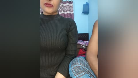 Video of a woman with medium skin tone, wearing a tight black sweater, sitting on a bed with a patterned blanket. Background shows a curtain and a blue bag hanging on a wall.