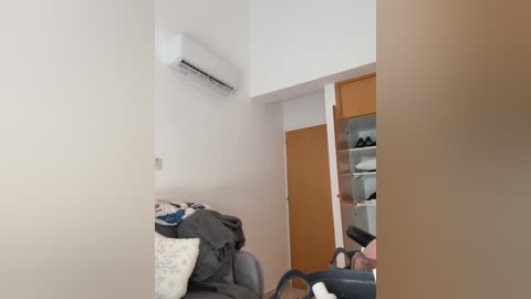 A video of a minimalist, modern bedroom with a white wall, an air conditioning unit, a partially open wardrobe revealing neatly organized clothes, and a gray couch with a white pillow.