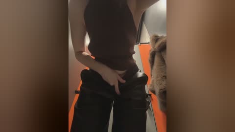 A video of a person in a dark, sleeveless top and black pants, adjusting their belt in a narrow, dimly lit hallway with orange walls and a brown coat hanging on a hook.