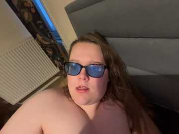 Video of a plus-sized, light-skinned woman with long brown hair, wearing blue-tinted glasses, lying on a gray bed, partially nude. Background includes a radiator and a colorful patterned curtain.