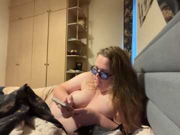 A video of a plus-sized, fair-skinned woman with long brown hair, wearing glasses, lying topless on a bed, reading a smartphone, in a cozy, cluttered bedroom.