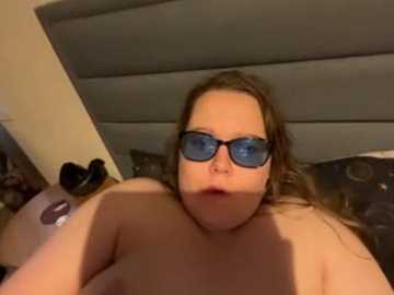 A video of a plus-sized, light-skinned woman with long brown hair, wearing black-framed glasses and lying topless on a bed. The background features a beige headboard and a patterned pillow.