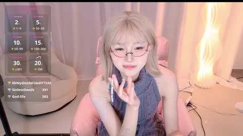 Video of a fair-skinned, petite, blonde-haired woman with glasses and a sleeveless top, sitting in a gaming chair with a cozy room background.