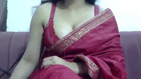 Video of a South Asian woman in a red sari with gold embroidery, sitting on a purple couch. The sari is partially draped, revealing her bare shoulders and cleavage. Background is a blurred indoor setting.