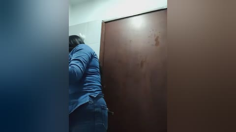 A video captures a person with short hair, wearing a blue shirt and jeans, bending over to open a brown wooden door, partially obscured by a blue curtain.